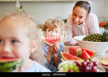 Donna felice che si diverte con il figlio che tiene una fetta di anguria fresca con frutta in cucina a casa Foto Stock