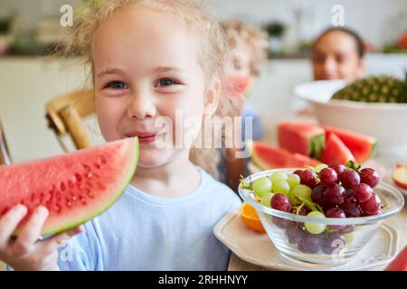 Graziosa ragazza bionda che sorride tenendo in mano l'anguria, fetta di uva in una ciotola a casa Foto Stock