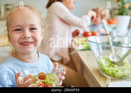 Ritratto di una ragazza bionda felice che tiene una ciotola di verdure mentre si siede con la famiglia a casa Foto Stock