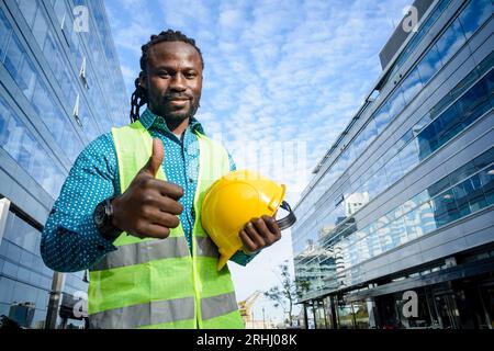 Felice, positivo giovane ingegnere civile di etnia africana con barba e dreadlocks, in piedi fuori dagli edifici commerciali, con berretto in han Foto Stock