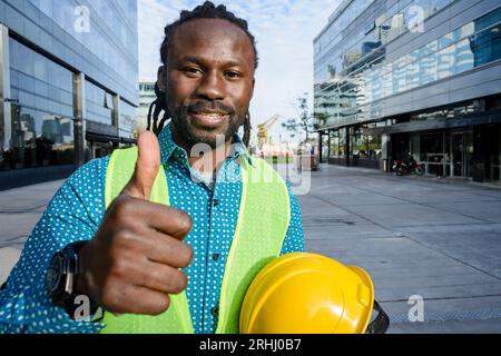 striscione di un giovane uomo nero positivo e felice con barba e dreadlocks ingegnere civile in piedi all'aperto con il pollice in alto, sorridendo e guardando la fotocamera w. Foto Stock