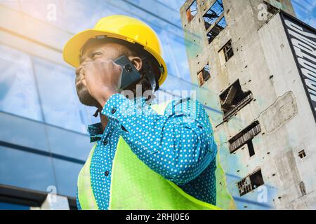 Doppia esposizione di un giovane ingegnere civile di etnia africana che indossa un elmetto di sicurezza giallo, in piedi all'aperto che parla al telefono con edifici in ba Foto Stock