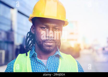 ritratto di un bell'ingegnere civile di etnia africana con barba e dreadlock, con elmetto di sicurezza giallo e giubbotto riflettente, è in piedi Foto Stock