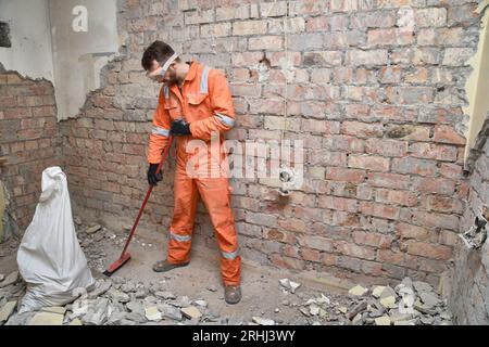 Stanza di pulizia per operai edili con scopa dopo lavori di demolizione, indossare guanti, maschere, stivali e tute arancioni. Foto Stock