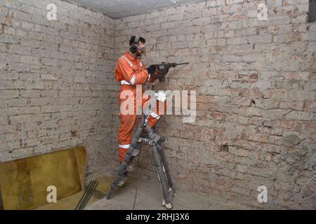 Operaio edile che rimuove il cemento vecchio dalla parete in mattoni all'interno, in piedi sulla scala utilizzando un trapano a martello, indossando guanti, maschere, cuffie antirumore e ora Foto Stock