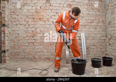 Costruttore che utilizza il miscelatore per impastare la soluzione di cemento nel secchio. Lavoratore che indossa DPI e tute arancioni. Foto Stock