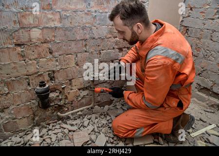 Un giovane idraulico maschio che perde collegamento con acqua, indossa guanti, stivali e tute arancioni. Foto Stock