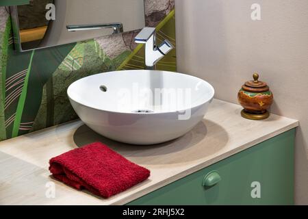 Interno del bagno con specchio riflettente fissato su pareti di piastrelle di design verde, mentre asciugamano rosso ripiegato posizionato vicino al moderno lavandino bianco con rubinetto e i Foto Stock