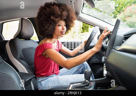 Vista laterale di una giovane donna afro-americana sorridente con i capelli afro che guarda lontano mentre si siede sul sedile del conducente e si imposta la direzione in satellite Foto Stock