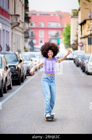 Corpo completo di allegra donna afroamericana in abiti casual con capelli afro che guardano la macchina fotografica mentre si guida sullo skateboard su strada asfaltata vicino al parcheggio c Foto Stock