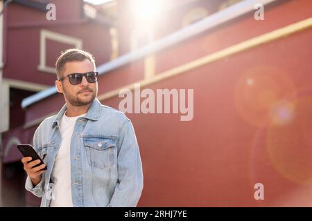 Bellissimo uomo che indossa una giacca in denim e occhiali da sole che cammina per la città. Foto Stock