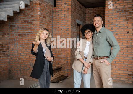 un broker immobiliare sorridente che punta con mano e mostra nuove abitazioni a coppie multiculturali Foto Stock