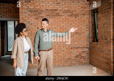 uomo felice che punta con la mano mentre mostra la nuova casa alla donna afroamericana Foto Stock