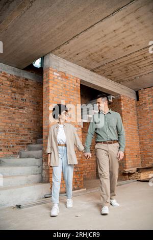 gioiosa coppia interrazziale che si tiene per mano e si guarda mentre cammina in un nuovo cottage Foto Stock