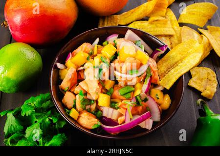 Ceviche di salmone con mango in una ciotola di legno con patatine di platana: Una ciotola di ceviche di salmone con patatine di platana circondata da ingredienti Foto Stock