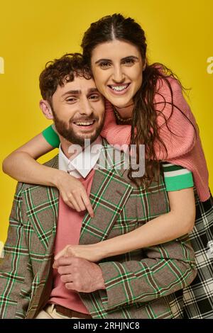 donna allegra che abbraccia un uomo barbuto, in blazer, sfondo giallo, abiti universitari, studenti felici Foto Stock