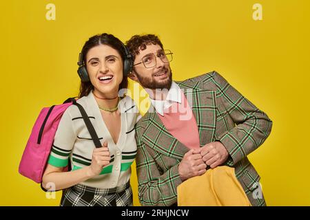 donna allegra con cuffie wireless in piedi vicino a un uomo barbuto, studenti che tengono zaini, musica Foto Stock