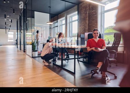 Un gruppo eterogeneo di professionisti aziendali collabora in un moderno centro di coworking startup, utilizzando un mix di strumenti cartacei e tecnologici Foto Stock