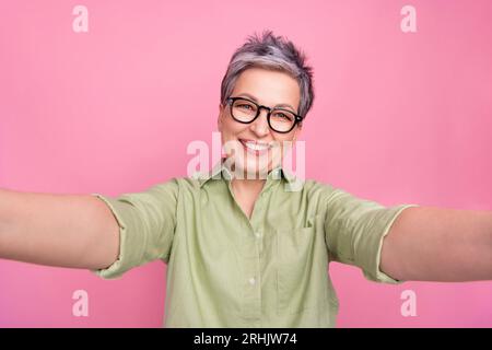 Primo piano selfie con i capelli grigi di una donna d'affari positiva di successo che registra il suo blog toothy sorridente isolato su sfondo rosa Foto Stock