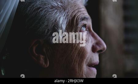 L'uomo anziano guarda da vicino il cielo dalla finestra di casa, osservando il tempo e la pioggia in arrivo. Persona anziana che fissa la formazione della tempesta Foto Stock