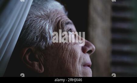 L'uomo anziano guarda da vicino il cielo dalla finestra di casa, osservando il tempo e la pioggia in arrivo. Persona anziana che fissa la formazione della tempesta Foto Stock