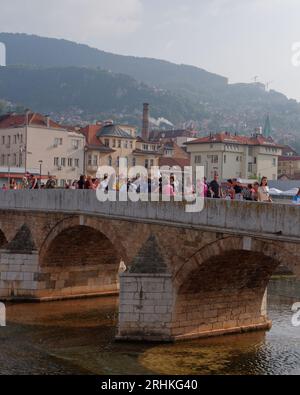 Persone su un ponte sul fiume Miljacka con il camino del birrificio Sarajevska pivara alle spalle. Sarajevo, Bosnia ed Erzegovina, 17 agosto 2023. Foto Stock