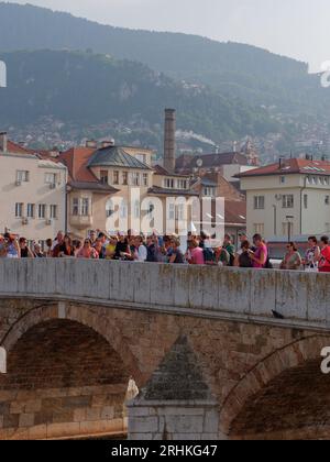 Persone su un ponte sul fiume Miljacka con il camino del birrificio Sarajevska pivara alle spalle. Sarajevo, Bosnia ed Erzegovina, 17 agosto 2023. Foto Stock