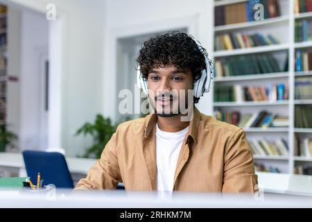 Un uomo serio e concentrato in cuffie che studia in biblioteca, che studia e registra un corso video mentre è seduto nella biblioteca del campus universitario. Foto Stock