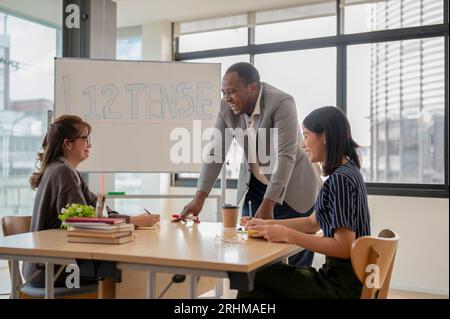 Un insegnante o tutor di inglese afroamericano professionista e allegro sta ridendo e godendo la conversazione con gli studenti asiatici durante la lezione. Foto Stock