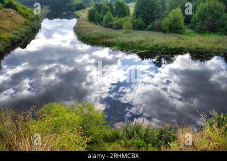 Sereno fiume circondato da vegetazione lussureggiante, l'acqua nel fiume è così placida che rispecchia le soffici nuvole bianche nel cielo sopra, dando un etereo Foto Stock