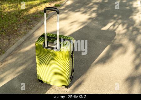 sul sentiero, inondato di luce solare, è presente una grande valigia verde su ruote e con manico telescopico. bagaglio da viaggio. Vacanza o viaggio d'affari. Foto Stock