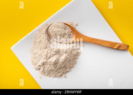 Polvere di farina di buccia di psicllium su piatto bianco e cucchiaio di legno in casa, sfondo giallo. Benefici per la salute del concetto di farina di Psyllium. Foto Stock