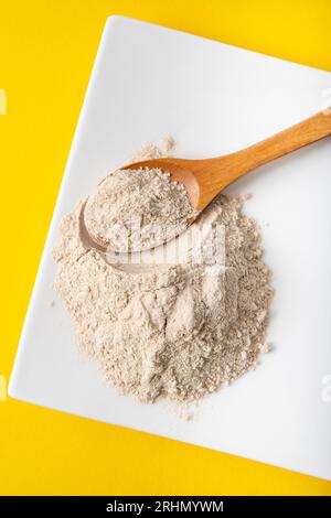 Polvere di farina di buccia di psicllium su piatto bianco e cucchiaio di legno in casa, sfondo giallo. Benefici per la salute del concetto di farina di Psyllium. Foto Stock