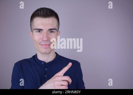 Sorridente e affascinante uomo caucasico con maglietta blu punta lateralmente su sfondo isolato Foto Stock