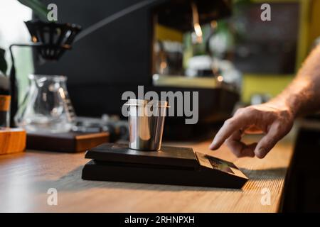 vista ritagliata del barista utilizzando bilance elettroniche e bicchiere vicino alla macchina per il caffè sfocato nella caffetteria Foto Stock