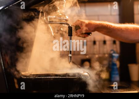 vista ridotta del barista sfocato utilizzando una macchina da caffè con vapore e illuminazione nella caffetteria Foto Stock