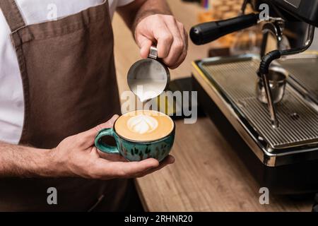 vista corta del barista nel grembiule che versa latte e prepara il cappuccino vicino alla macchina del caffè nella caffetteria Foto Stock