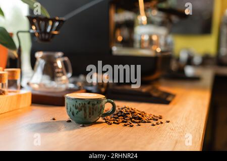 tazza di cappuccino vicino ai chicchi di caffè sul piano di lavoro vicino alla macchina da caffè sfocata nel caffè Foto Stock