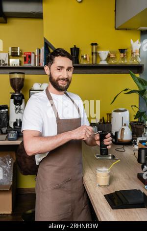 allegro barista che si occupa della macinazione del caffè al grembiule e che guarda la macchina fotografica vicino a una bevanda fredda al caffè nel bar Foto Stock