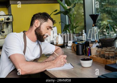 barista barbuto in grembiule che scrive sul notebook vicino alla tazza e ai chicchi di caffè in caffetteria Foto Stock