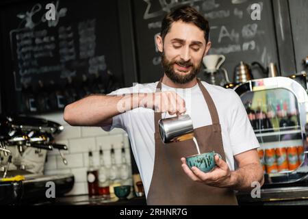 barista positivo nel grembiule che versa latte dalla caraffa in tazza con cappuccino in caffetteria Foto Stock