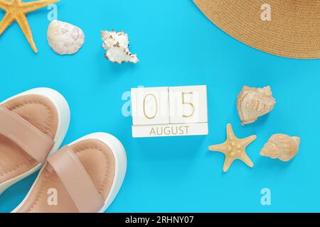 Agosto 05. Calendario in legno su sfondo blu con accessori estivi, vista dall'alto. Vacanze estive. Calendario perpetuo, stivali, cappelli e conchiglie con Foto Stock
