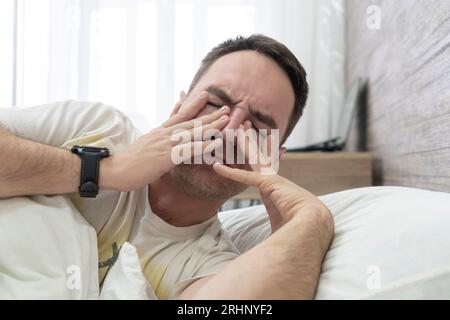 Fatigue Concept. Tired Young Man In Bed At Home. Exhausted Millennial Guy Waking Up In Bedroom . Feeling Unwell, Having Headache Or Suffering Insomnia Stock Photo