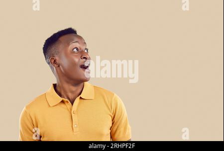 Il giovane uomo afro-americano WOW positivo guarda in alto con emozioni entusiastiche si trova nello studio beige Foto Stock
