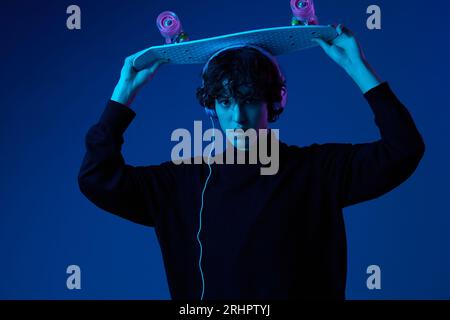 Adolescente maschio con cuffie che ascoltano musica e danza skateboard in mano sulla sua testa, stile di vita hipster, ritratto sfondo blu scuro, neon Foto Stock