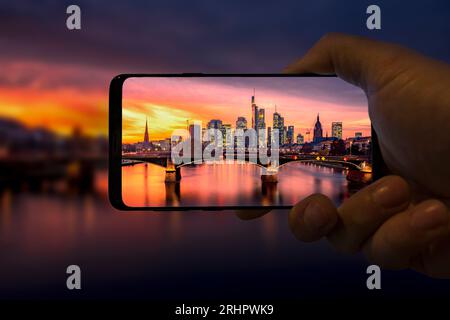 Smartphone in mano mostra lo skyline di Francoforte sul meno al tramonto, romantico bagliore serale, grattacieli illuminati, Assia, Germania Foto Stock