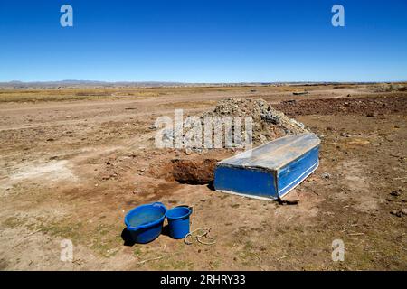 Lago Titicaca, BOLIVIA; 18 agosto 2023: Una barca a remi in legno ribaltata offre ombra per un pozzo scavato dagli abitanti del villaggio sulla riva secca della baia di Cohana sul lago interno / Huiñay Marka (la parte più piccola del lago Titicaca) vicino al villaggio di Cohana. I livelli dell'acqua nel lago Titicaca si stanno avvicinando al livello minimo record stabilito nel 1996, il più basso da quando il servizio meteorologico della Bolivia (Senhami) ha iniziato a tenere registri nel 1974. Molti incolpano il cambiamento climatico; gli ultimi anni sono stati più secchi del normale e El Niño si sta attualmente rafforzando nell'Oceano Pacifico al largo del Sud America. Foto Stock