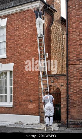 Pittore di case all'esterno che lavora su una scala per raggiungere il tetto con un collega in piedi sulla scala per evitare che scivoli Inghilterra Regno Unito Foto Stock