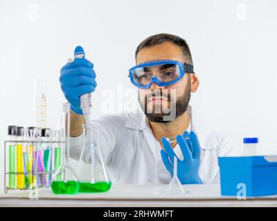 Giovane scienziato maschio che indossa occhiali protettivi per prelevare un campione di liquido da un bicchiere utilizzando una micropipetta Foto Stock