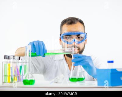 Giovane scienziato maschio che indossa occhiali protettivi in laboratorio versando un campione liquido da un cilindro graduato in un becher Foto Stock